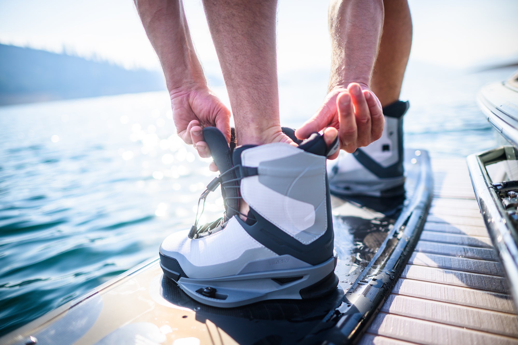 Person tightening wakeboard boots on a board near the water.