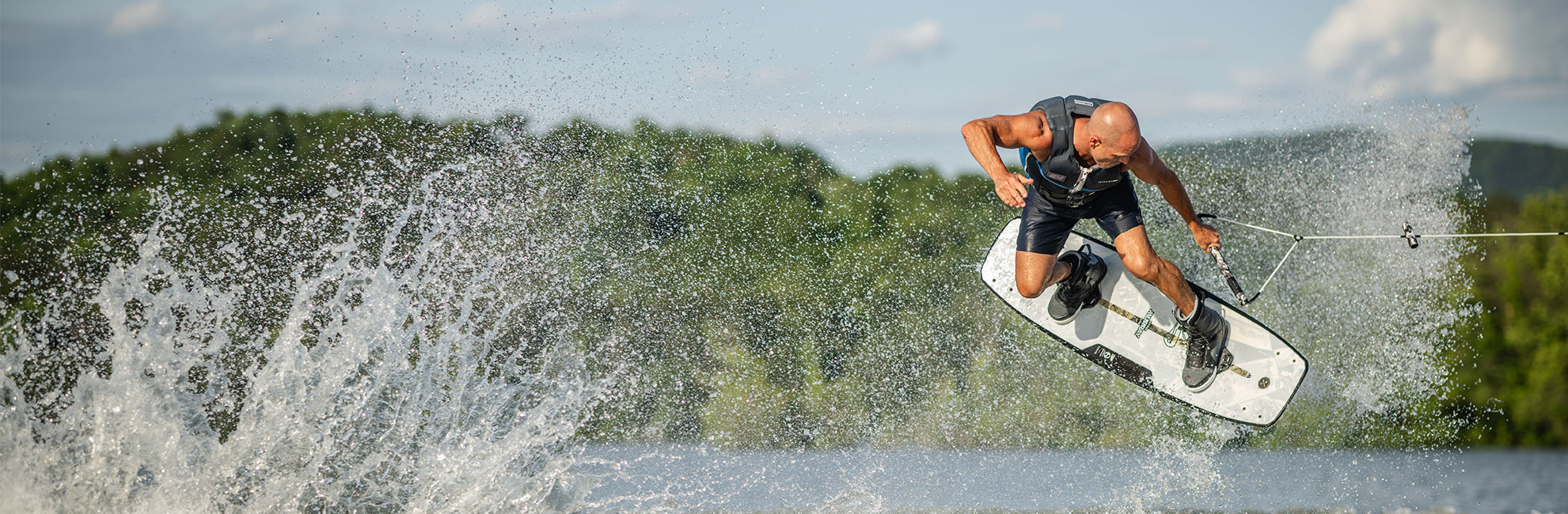 Person wakeboarding, performing a jump over splashing water, with a lush, green forest in the background.